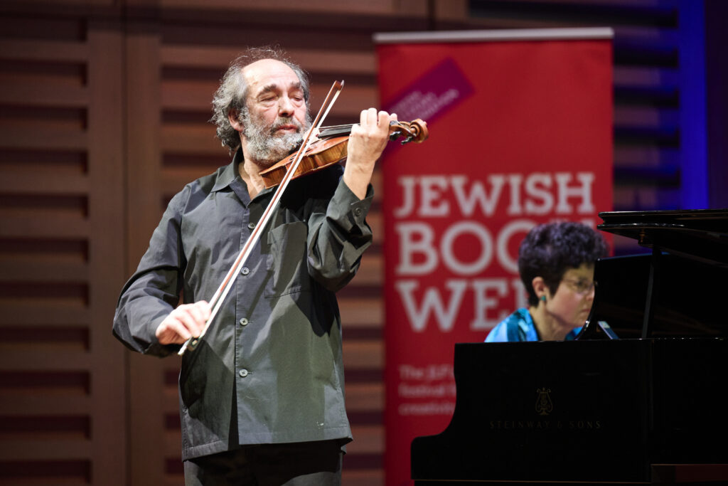 A man plays a violin on stage. Behind him, a banner reads Jewish Book Week and a woman sits with her body obscured by a grand piano.